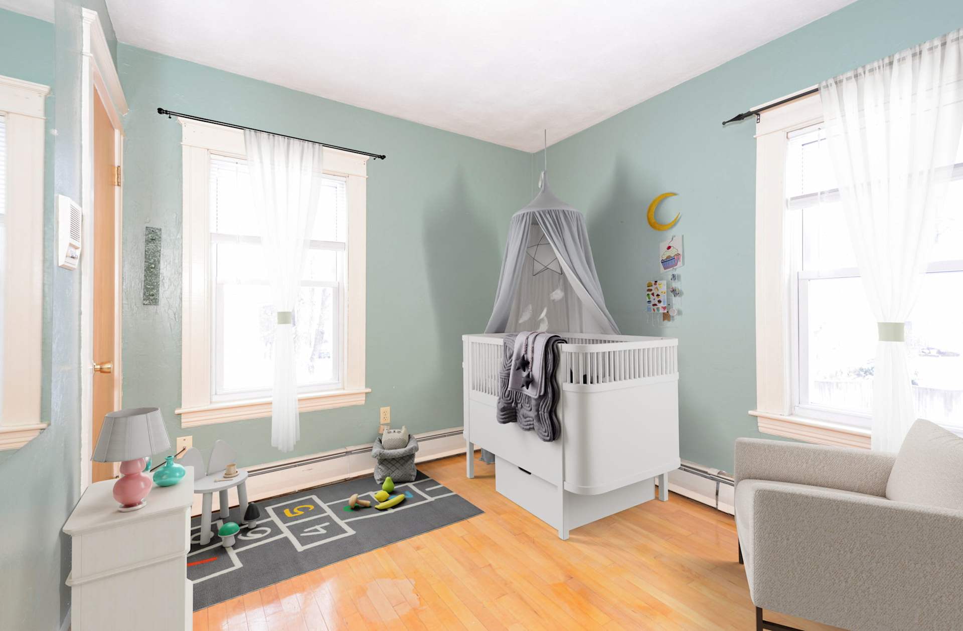 Nursery with a white canopy crib, mint-green walls, large windows, and a hopscotch rug.