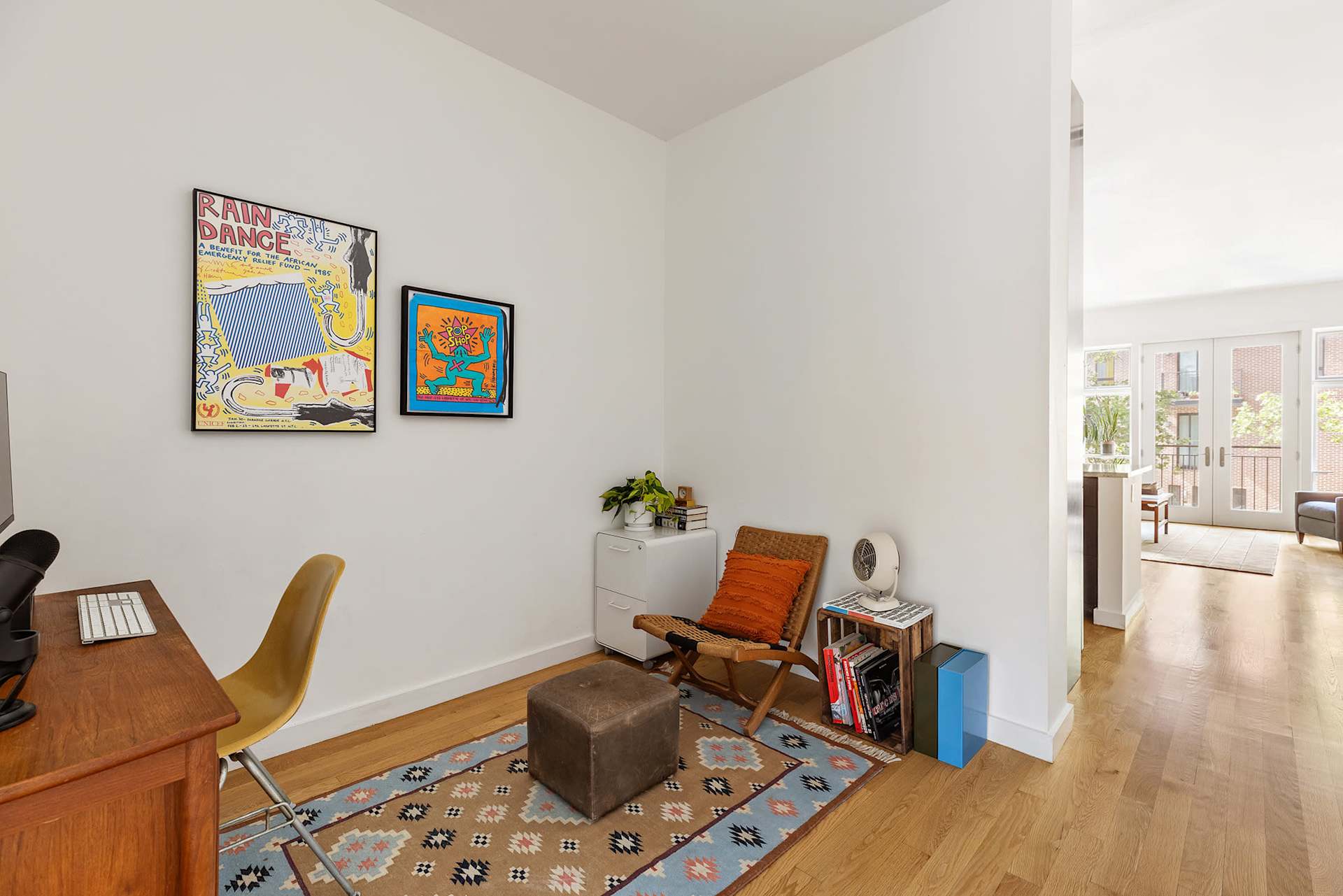 Home office nook with a wooden desk, colorful framed posters, and a woven chair beside the bright living area.