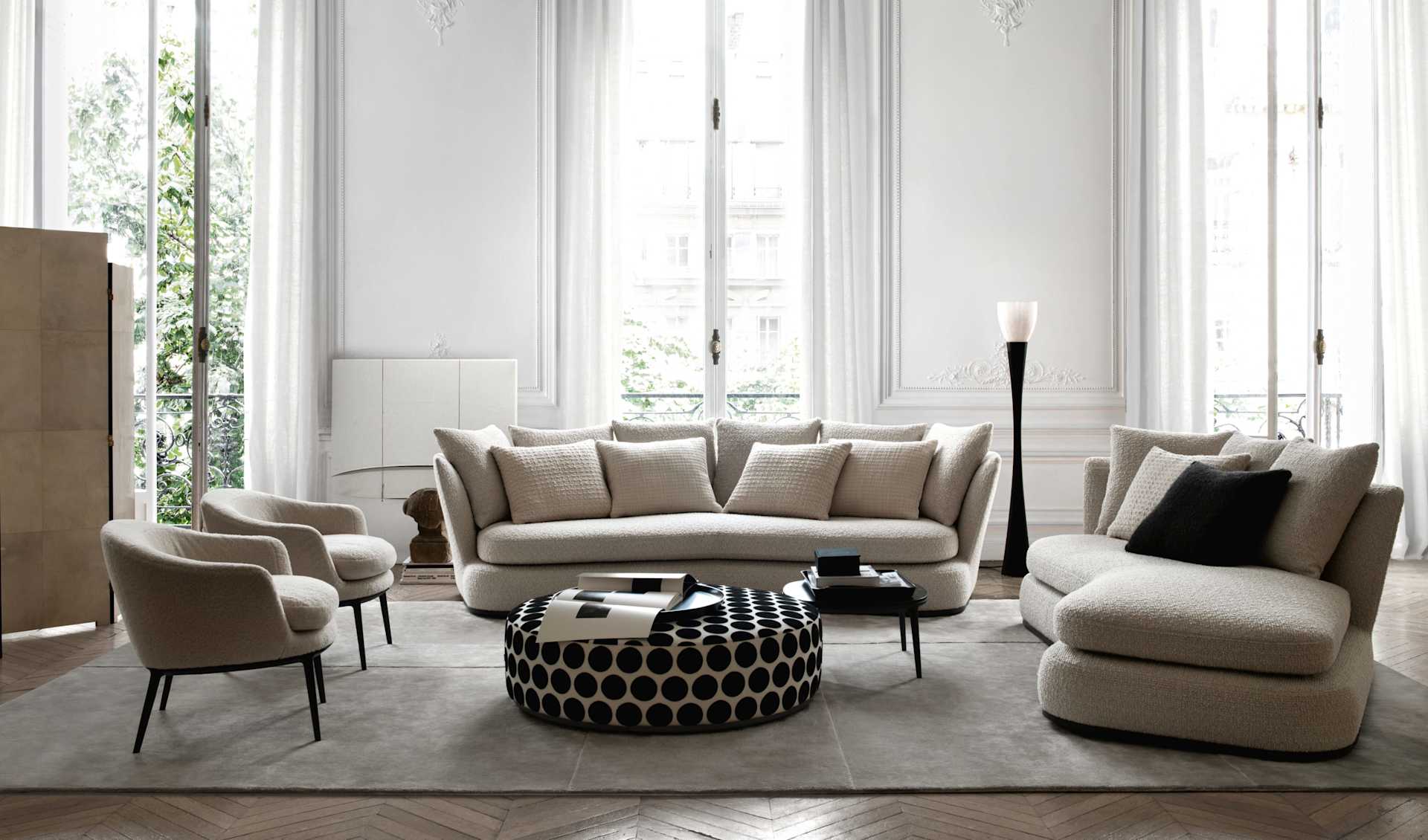 Bright living room with cream seating, tall windows, ornate molding, and a patterned round ottoman.