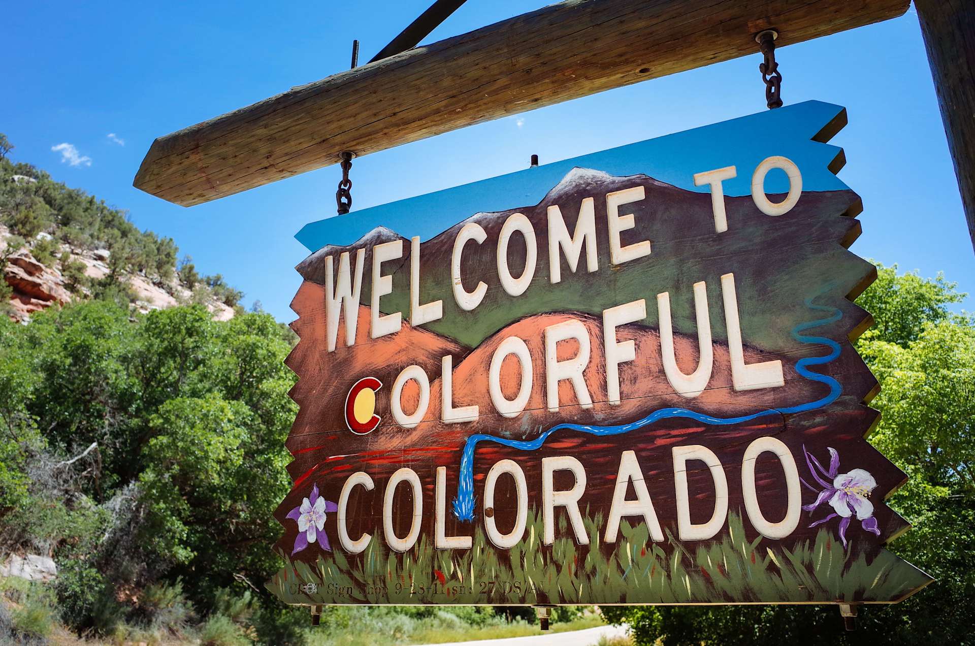 Hand-painted Welcome to Colorado sign beside a road, surrounded by trees and rocky terrain.