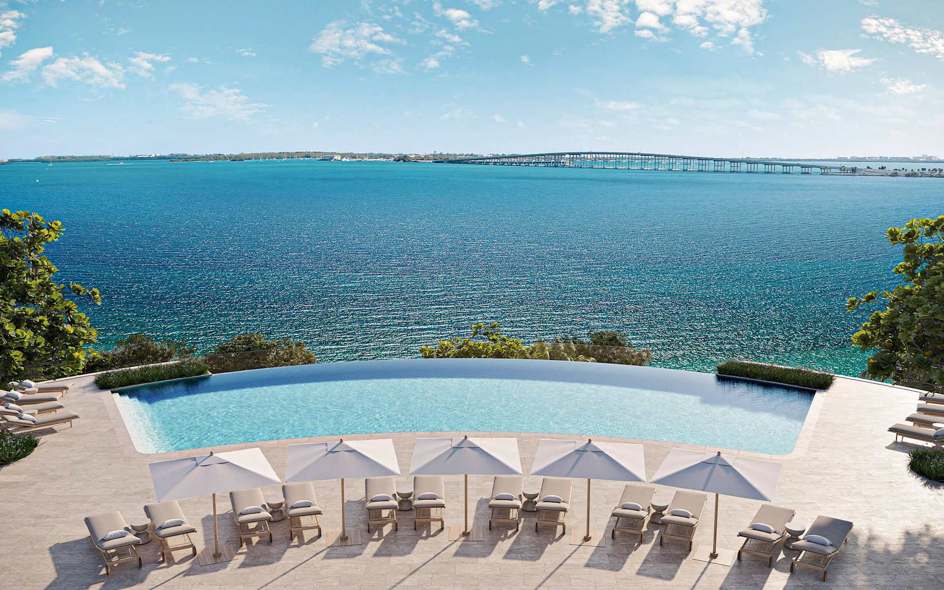 Pool terrace overlooking the bay, with rows of shaded lounge chairs and a bridge in the distance.
