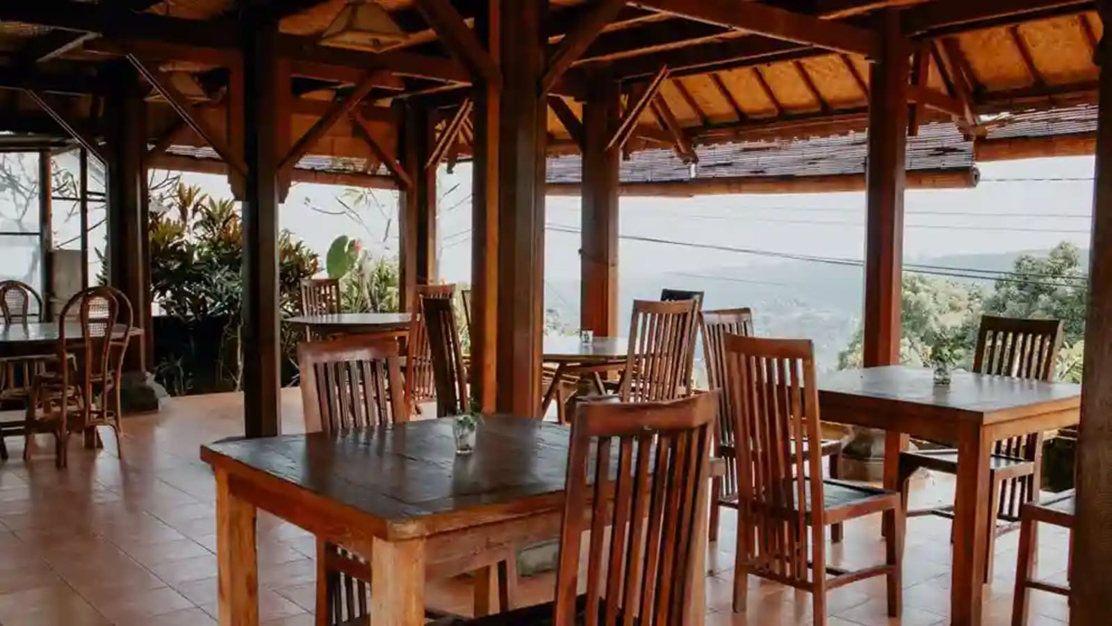 Spacious open-air Balinese dining verandah with solid wood tables, rattan hanging lanterns, guests dining happily overlooking rice terraces and mountain hills in Munduk Bali