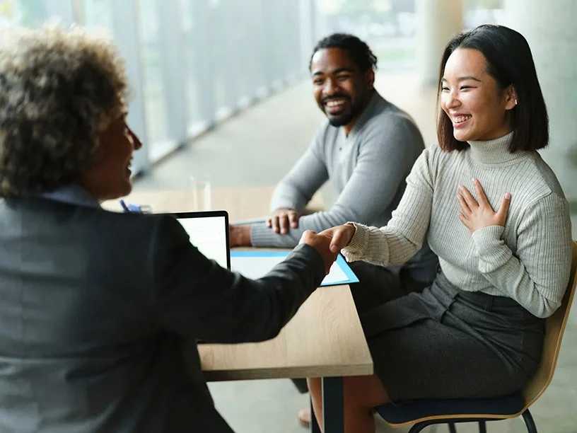 A financial service employee in a jacket assists two customers at a table with a laptop computer open, they are shaking hands and the customers are smiling, financial paperwork is on the table