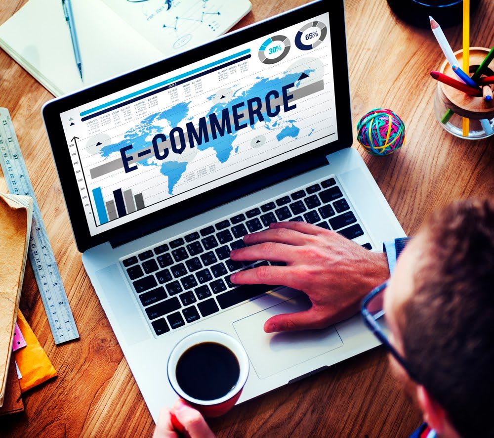 A laptop screen with the word ecommerce on it. An overhead POV of a man with coffee and his hand on the keyboard.