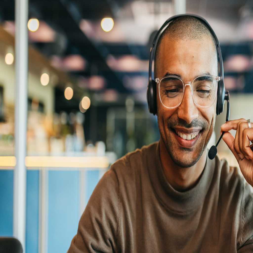 an education web hosting support team member in a headset is in glasses, he smiles and speaks into his headset while lightly gripping the microphone with his fingers