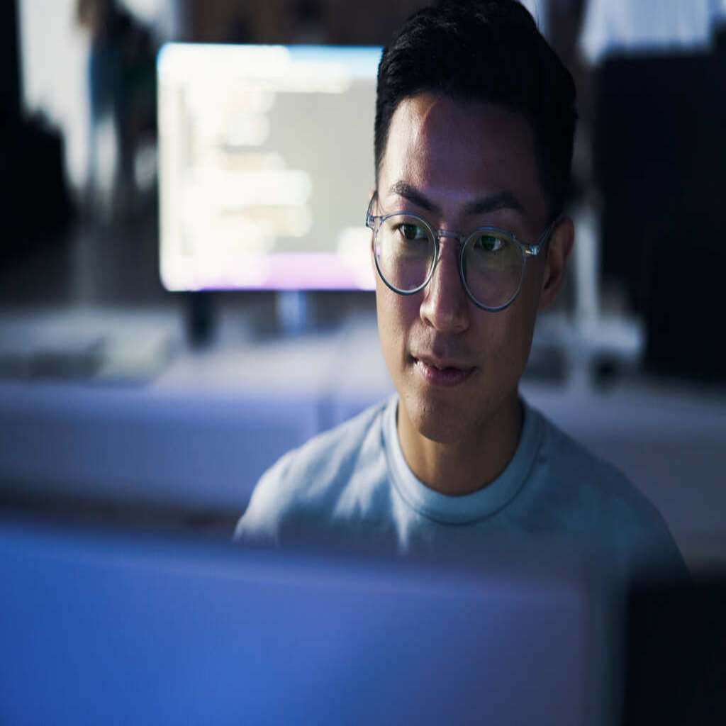 A young man in glasses working on building a hosted WooCommerce store, a computer at a desk behind him shows computer code