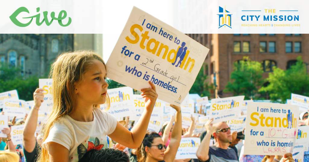 A young girl sits on the shoulders of an adult. She has blond hair and a somber look on her face. She holds up a sign that says, "I am here to stand in for 2nd grade girl who is homeless." At the top left, there is a Give logo. Top right, there is a logo for The City Mission.
