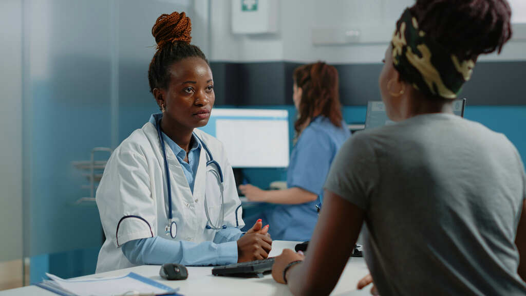 A female doctor stands behind a desk at a medical facility speaking to a patient.