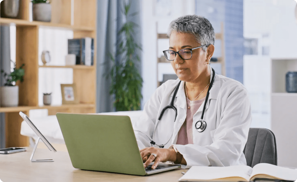 A telehealth doctor with a stethoscope around her neck works at her laptop.