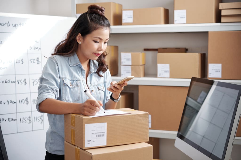 Woman packing product boxes for her business, looking at her cell phone to confirm information, ecommerce business models