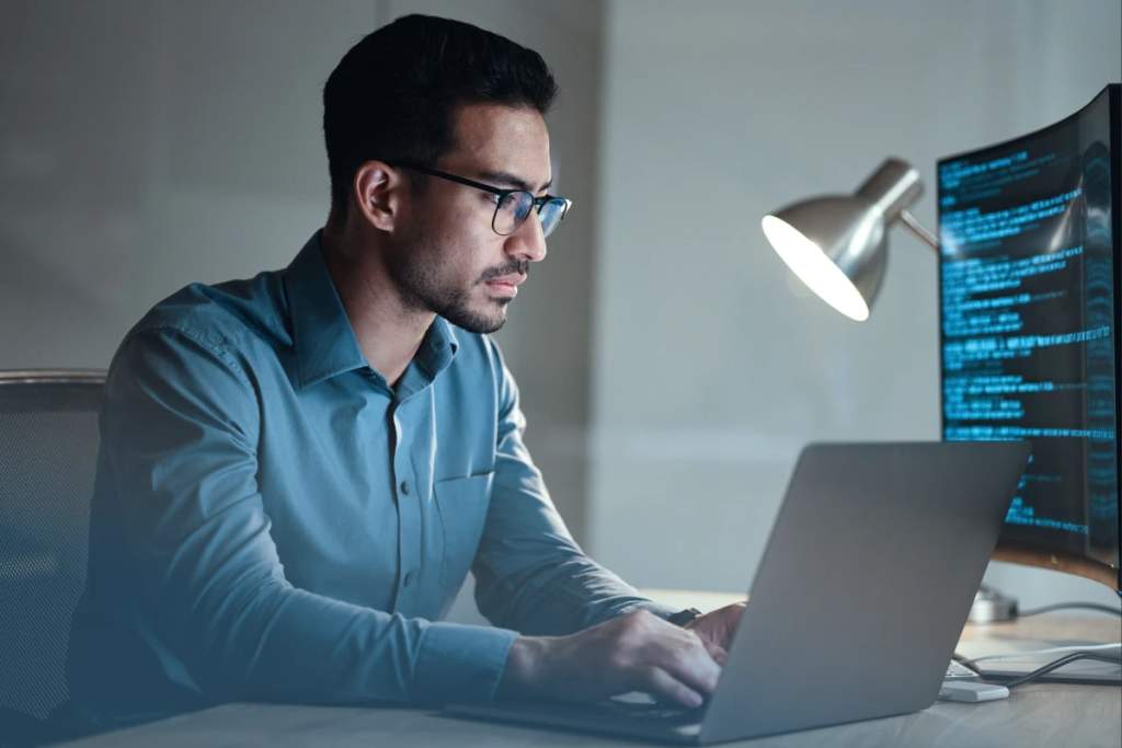 Man in dress shirt and glasses sits at desk in front of laptop and multiple monitors