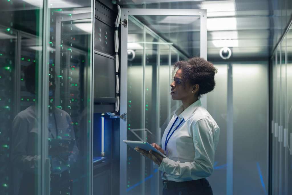 Woman in a blouse stands in a server room with a clip board