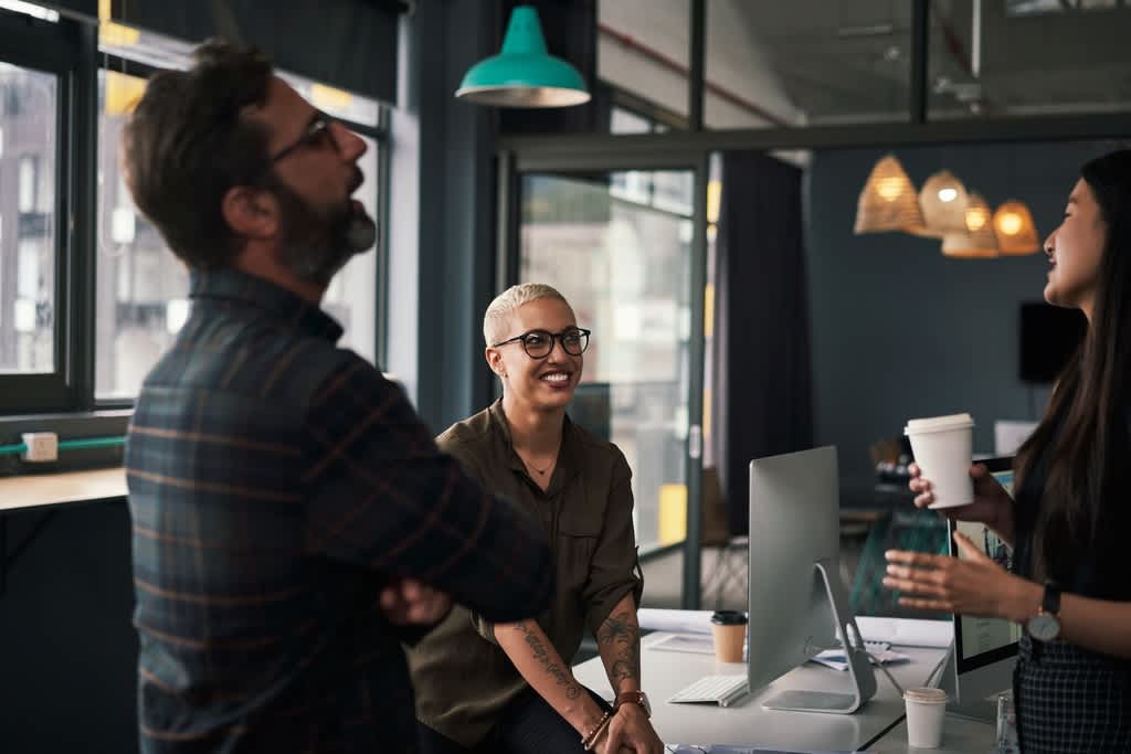 Three coworkers, two women and one man, in a break room with coffee, laughing with ease