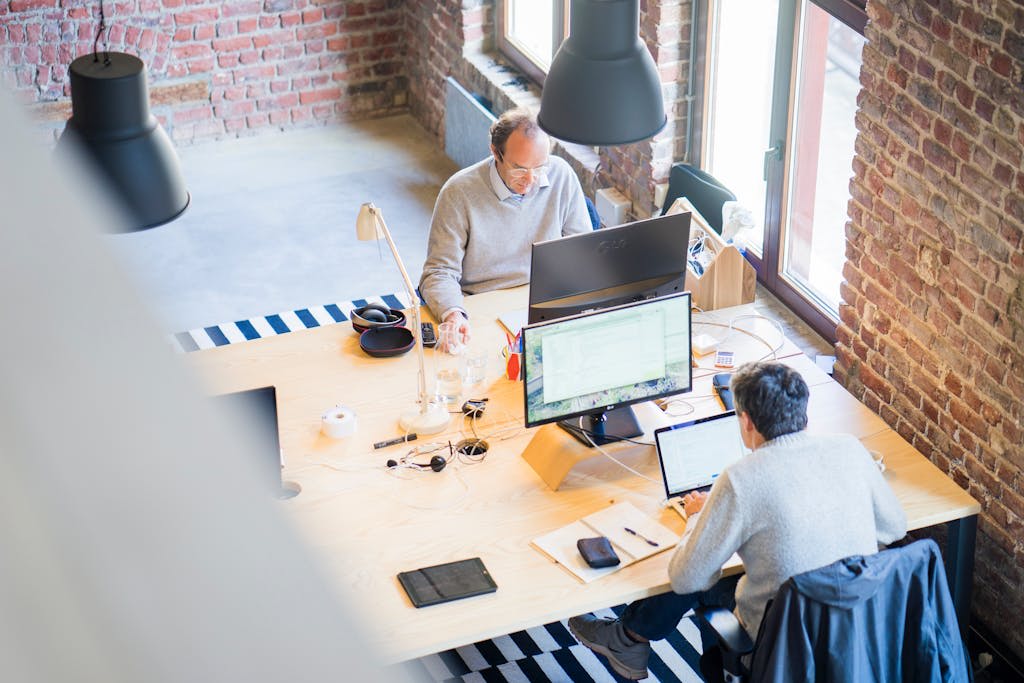 Two IT Experts Sitting While Facing Each Other on Flat Screen Monitors In Brick Coworking Space with Cement Floors
