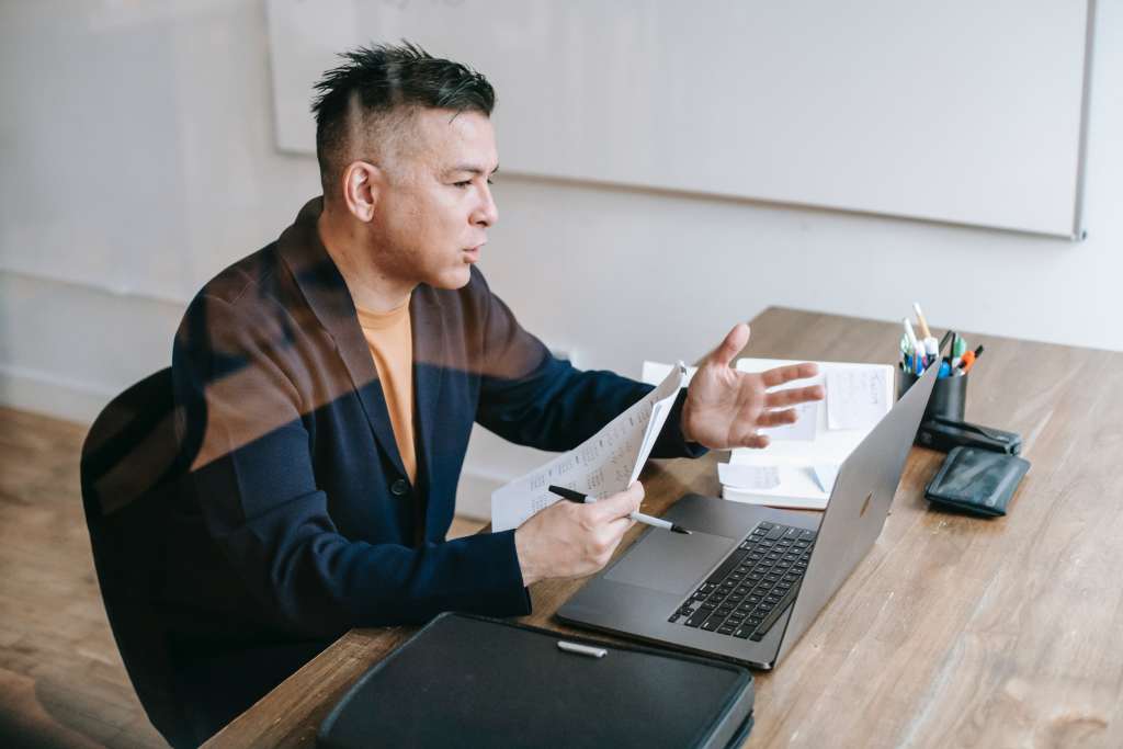 a man with short hair and a suit jacket holds an invoice and a pen while he talks on a video call