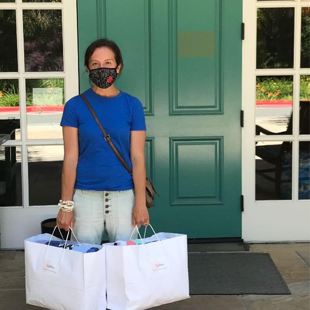 Liz, masked, standing in front of GMCH with two large bags in her hands, each stuffed with a quilt.