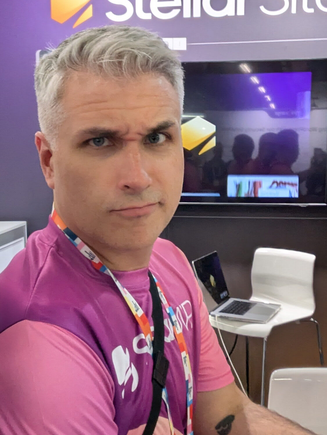 A person with gray hair and a skeptical expression wearing a purple shirt and a lanyard, standing in front of a booth with "Stellar" branding.