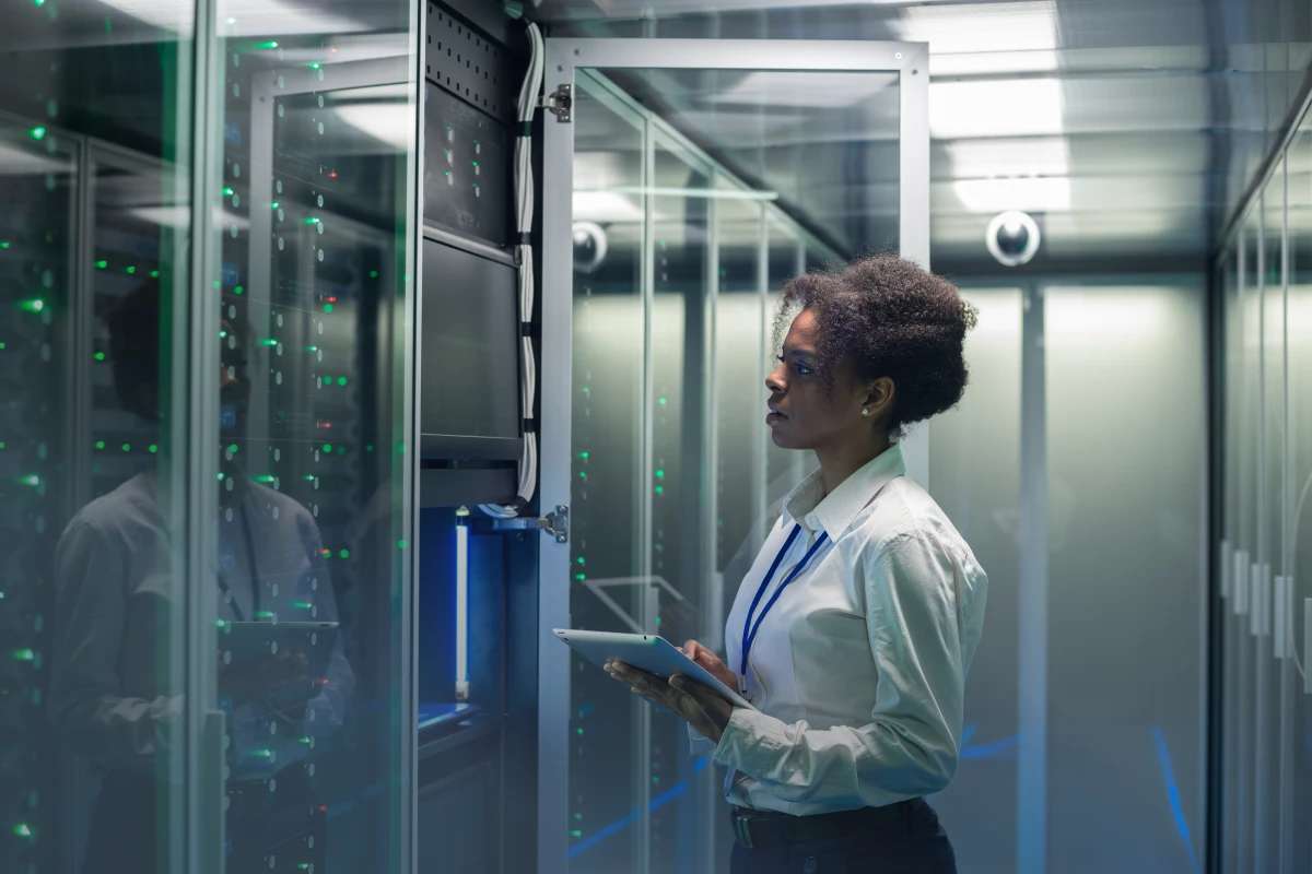 Woman in a blouse stands in a server room with a clip board