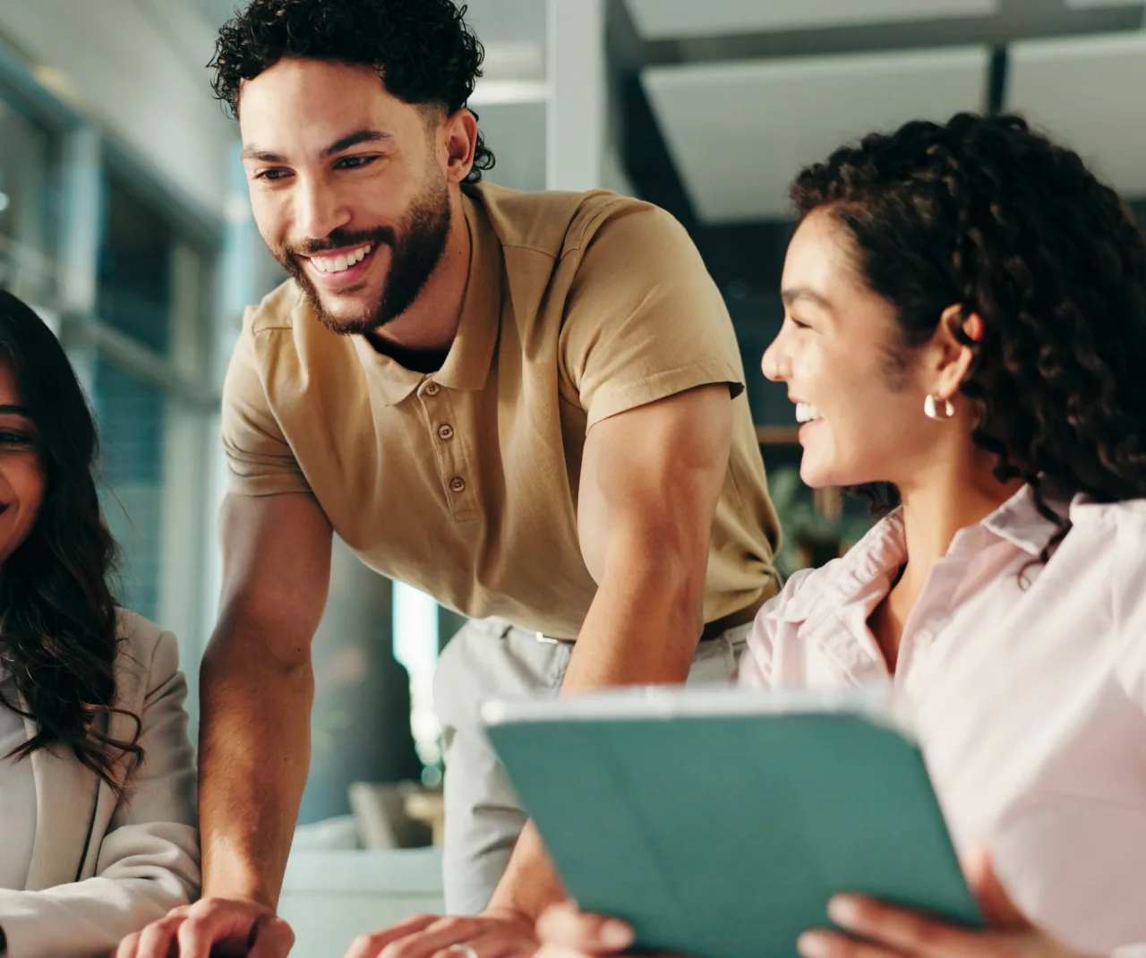 A man leans on a desk in an office smiling, his coworker's head is turned as she explores web hosting options on here tablet.