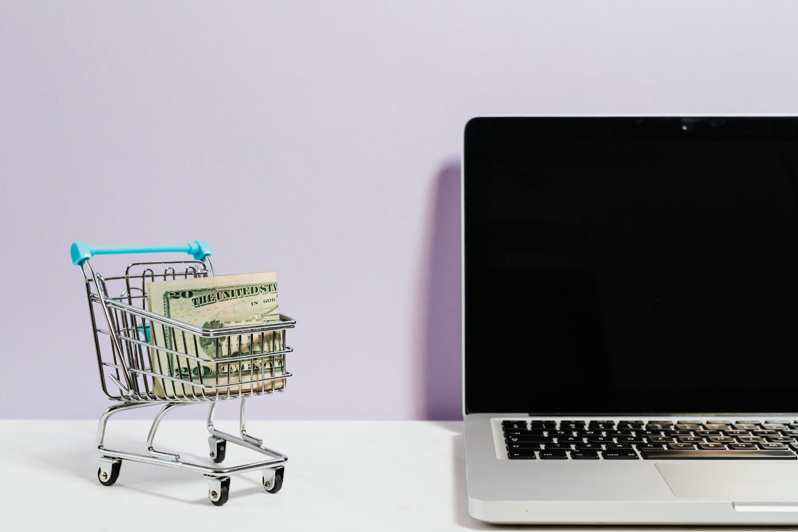 Tiny cart with money next to a laptop.
