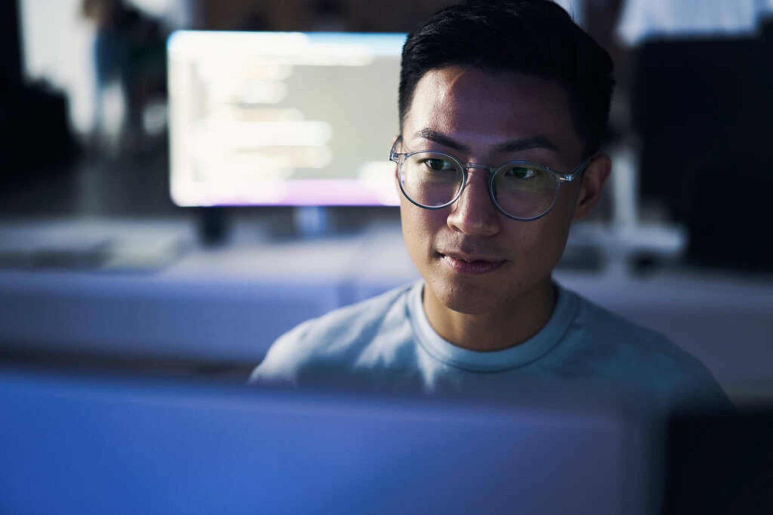 A young man in glasses working on building a hosted WooCommerce store, a computer at a desk behind him shows computer code