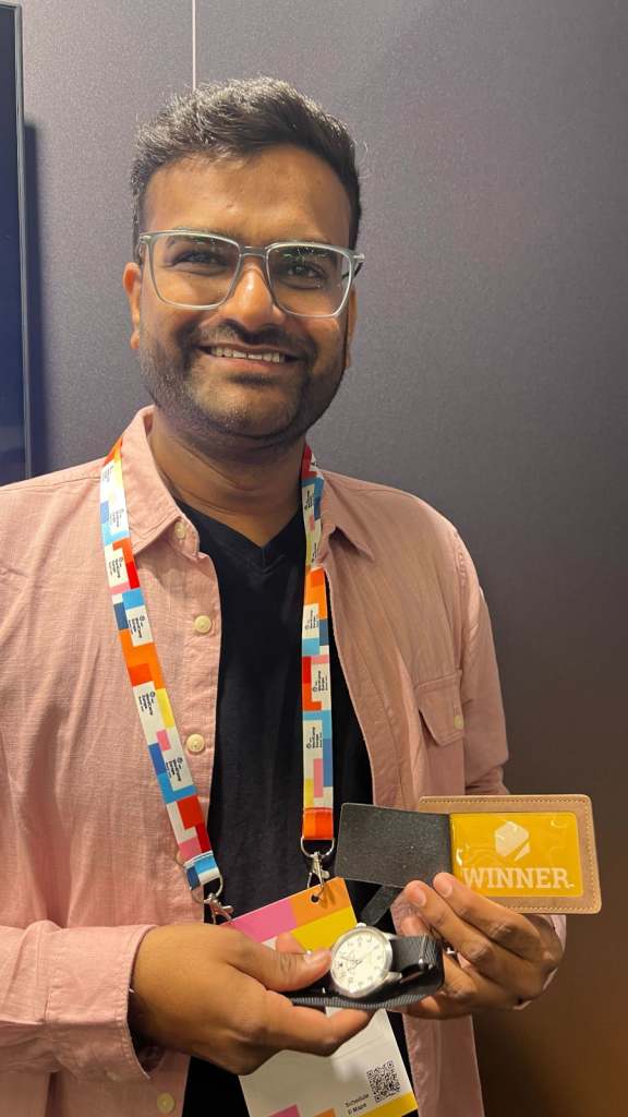 A man wearing glasses and a pink shirt, holding a watch and a card labeled 'Winner', with a colorful lanyard around his neck.