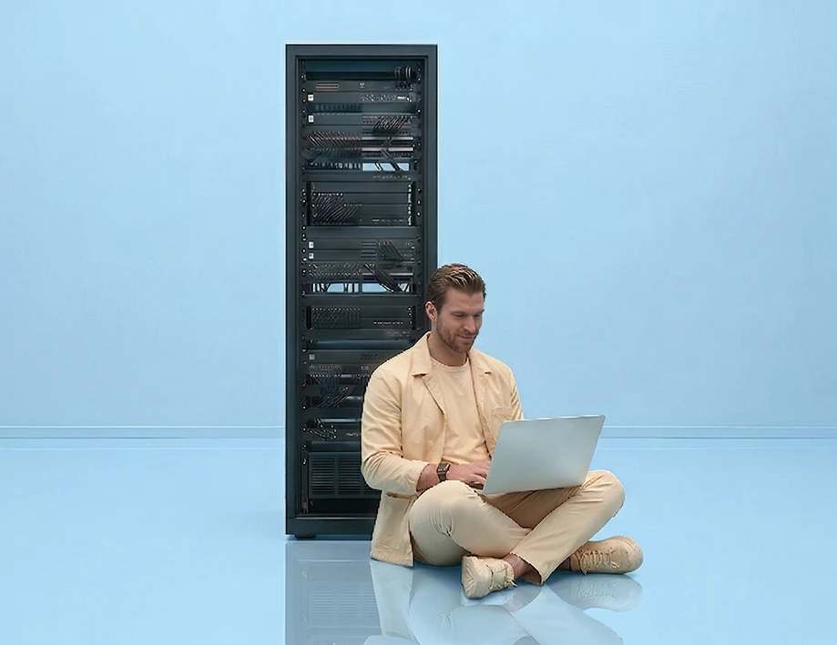 A man works on a laptop next to a server cabinet.