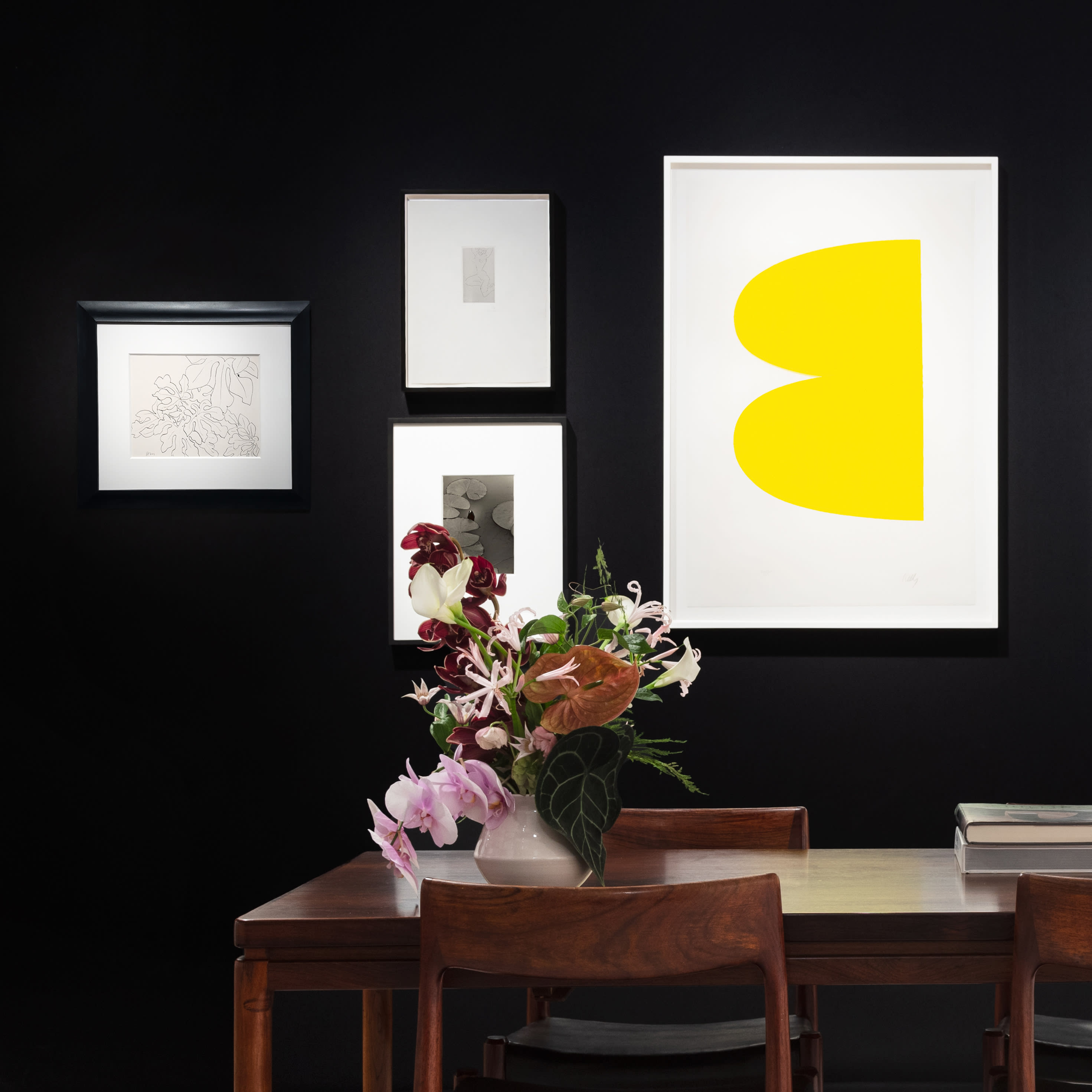 Installation view at Salon Art + Design 2024 in New York, with framed works clustered on a black wall above a table with chairs, books, and flowers.