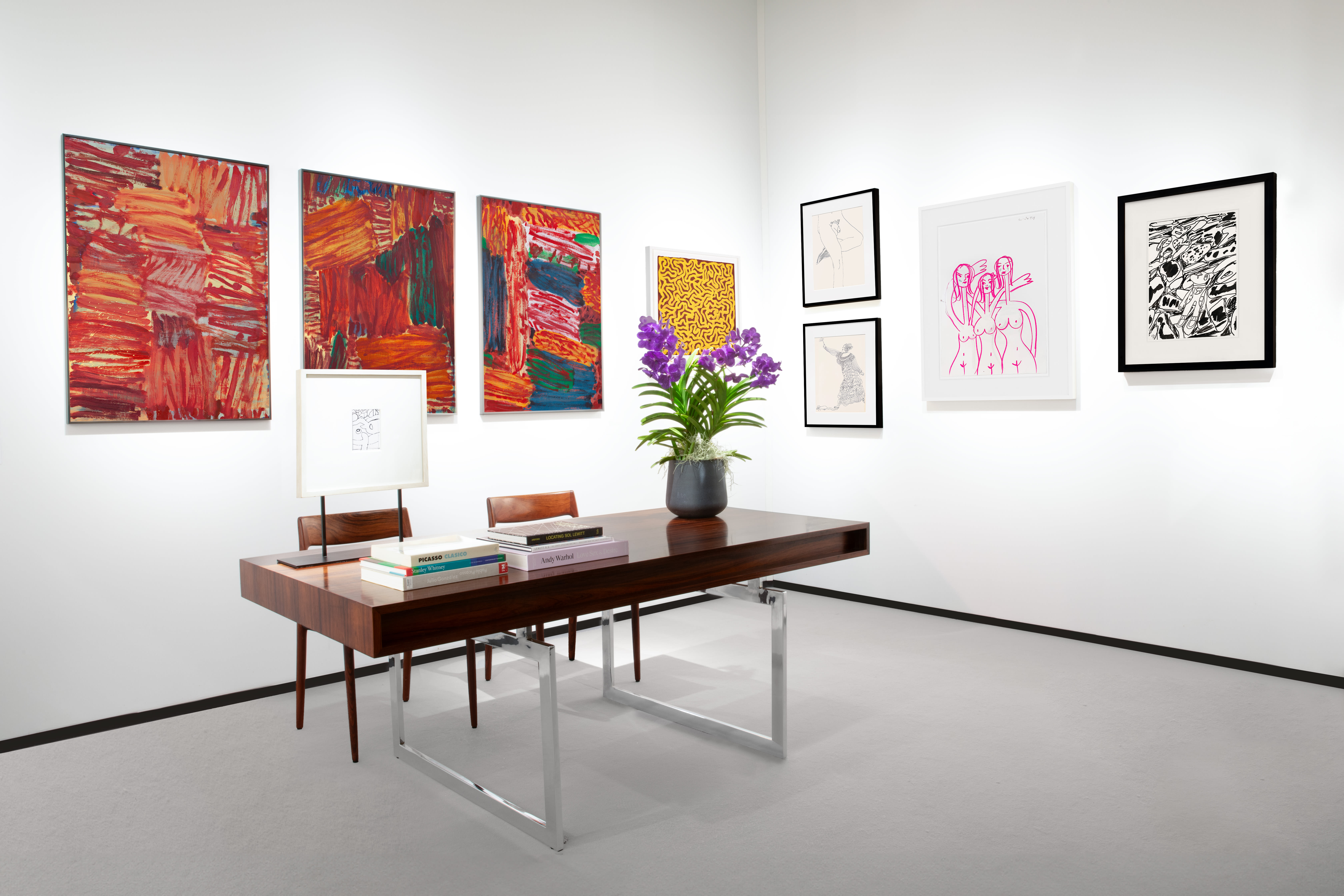 Installation view at Salon du dessin 2024 in Paris, with framed works across two corner walls above a table with flowers, books, and chairs.