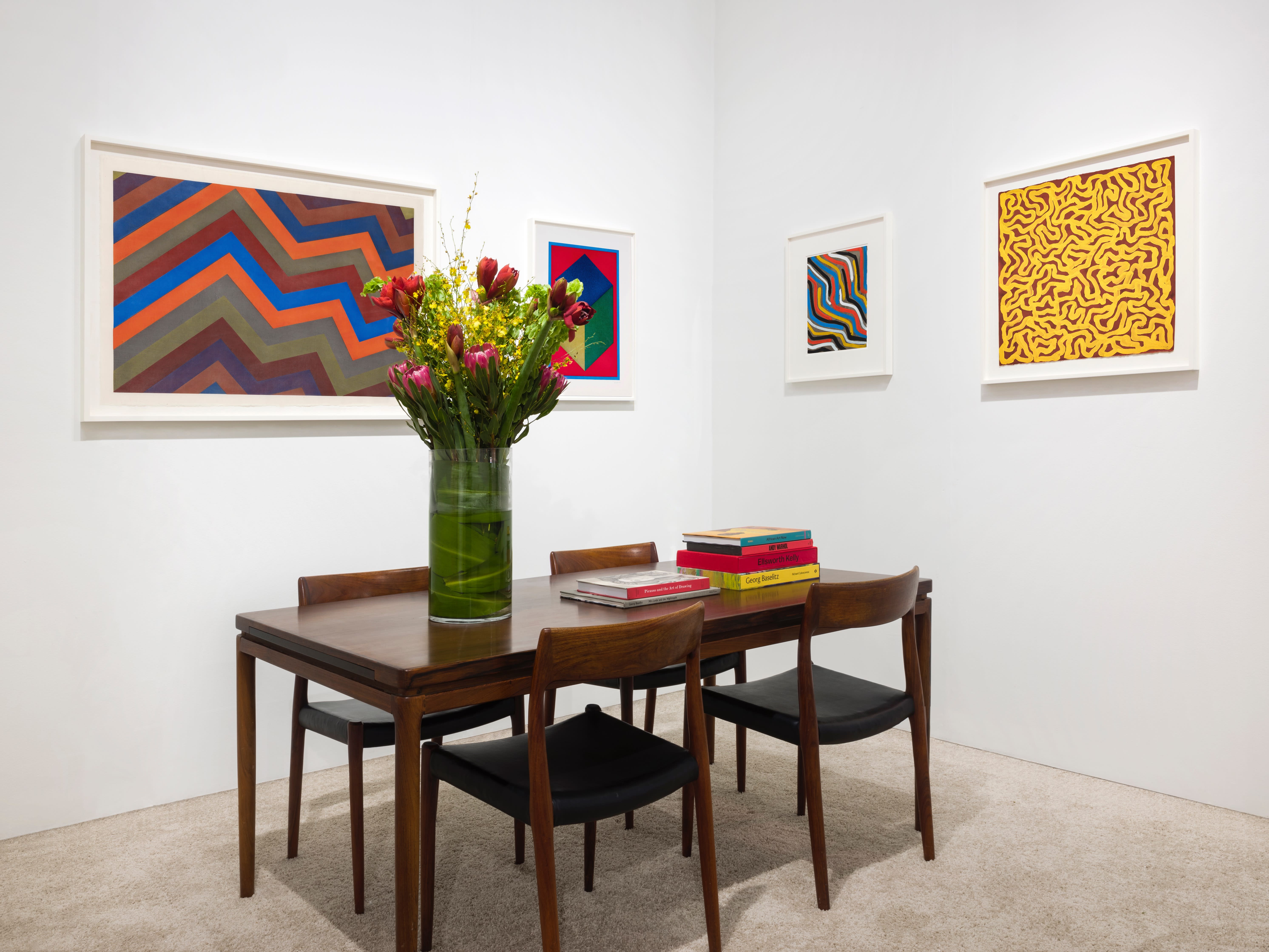 Installation view at Art Miami 2023 in Miami, with framed works across two corner walls above a table with chairs, books, and flowers.