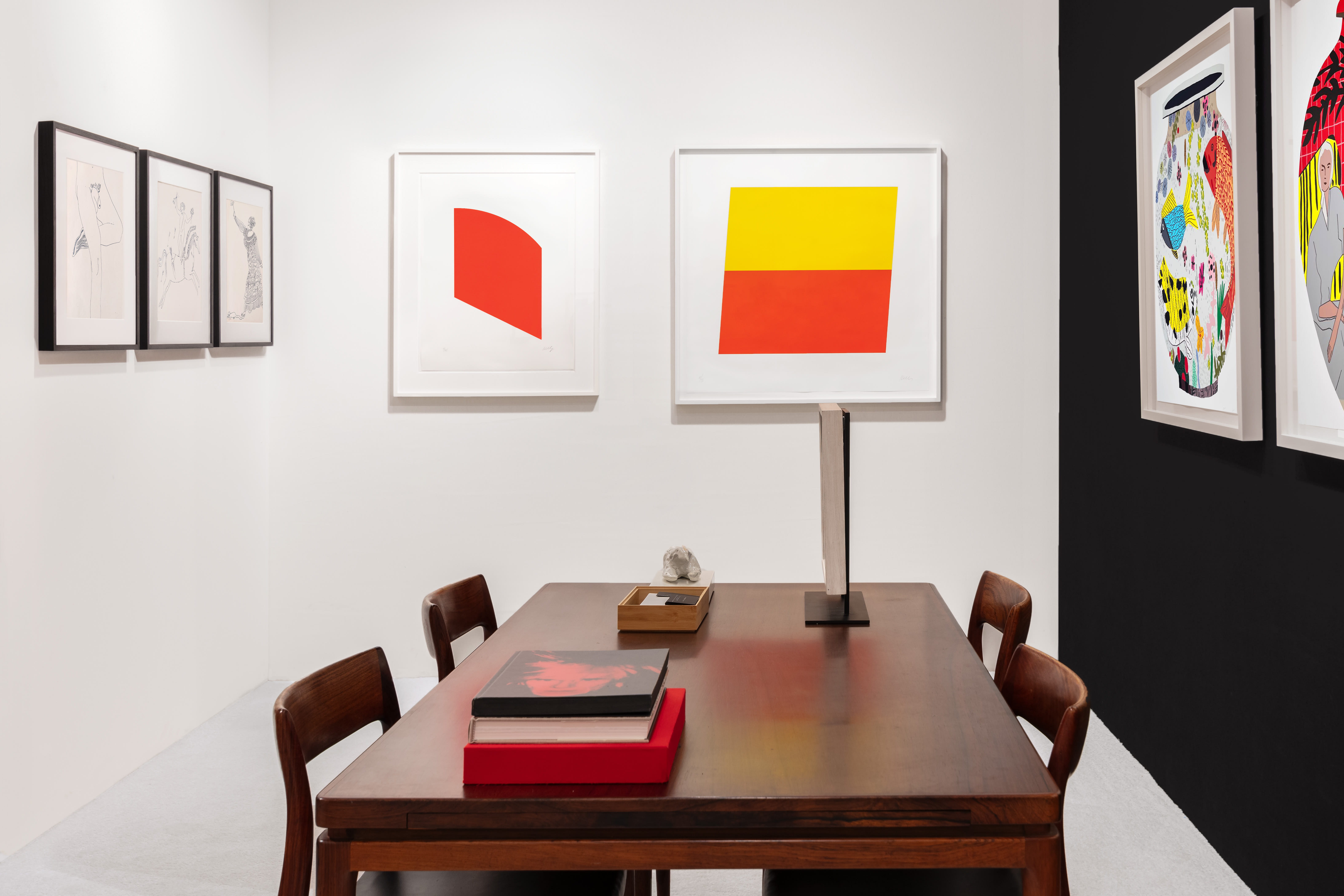 Installation view at Salon Art + Design 2023 in New York, with framed works across a white wall and adjacent black accent wall above a table with books and chairs.