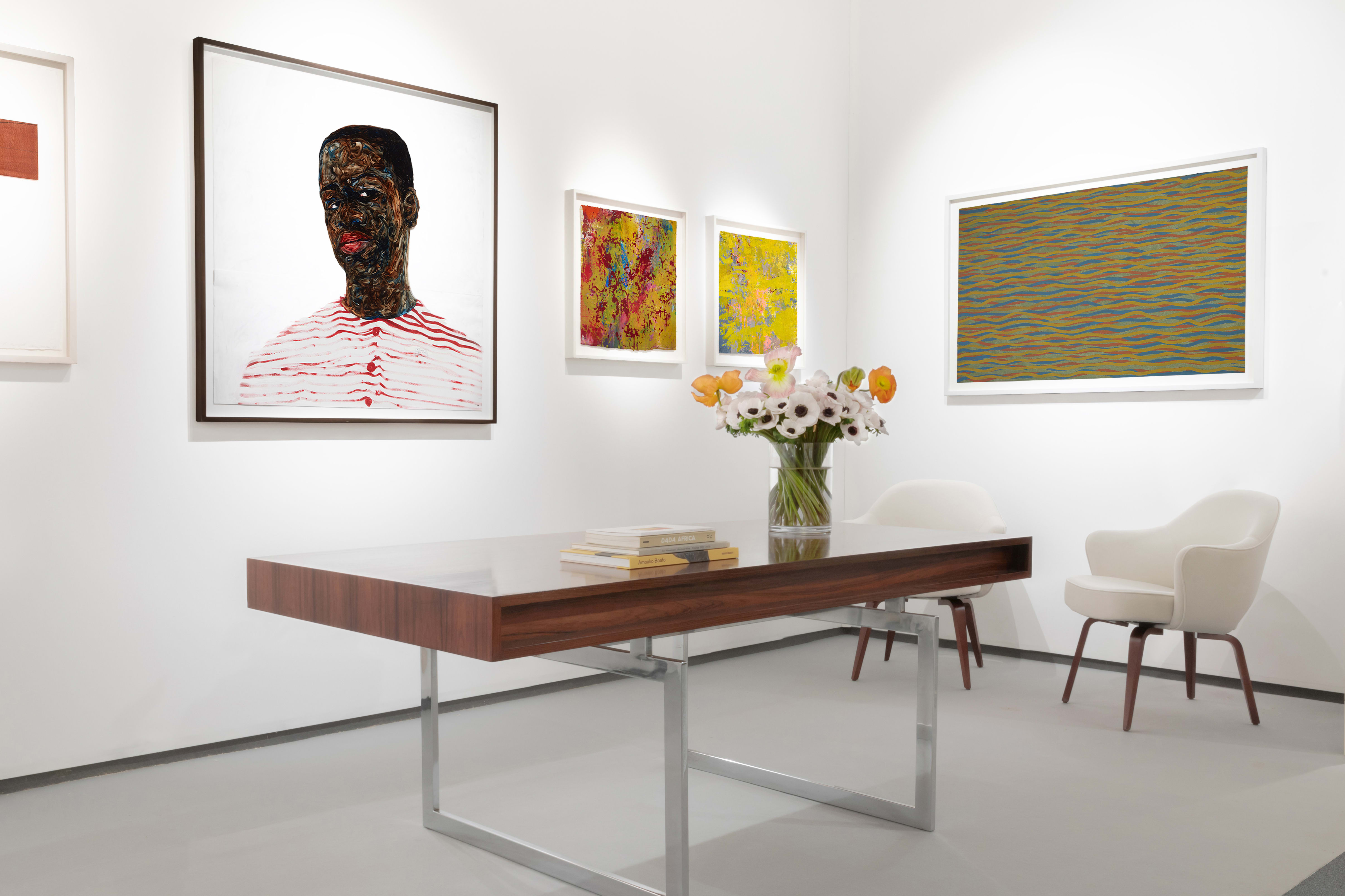 Installation view at Salon du dessin 2023 in Paris, with framed works across two corner walls above a table with flowers, books, and chairs.