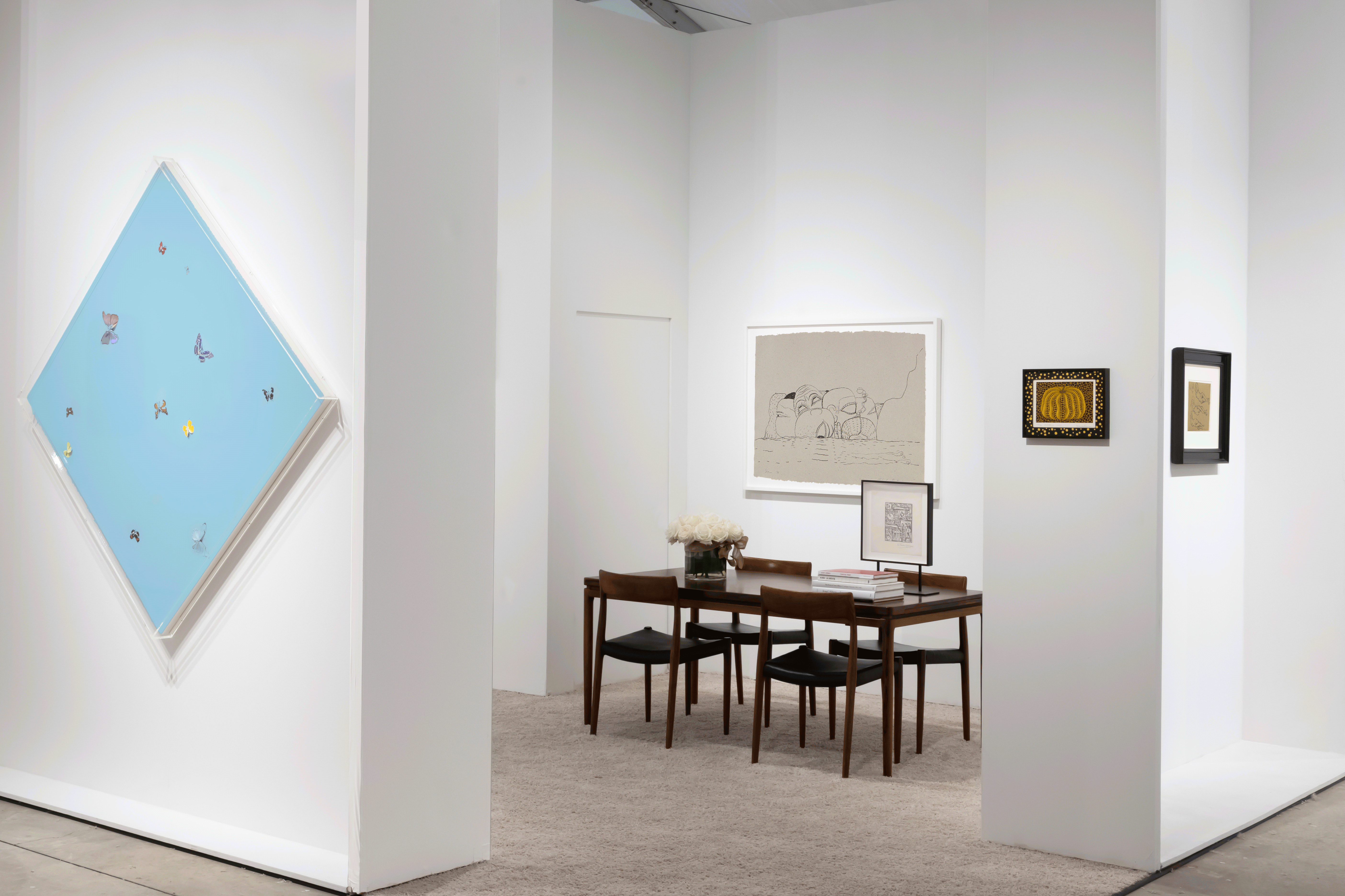 Installation view at Art Miami 2022 in Miami, with framed works across two corner walls above a table with chairs, flowers, and books.