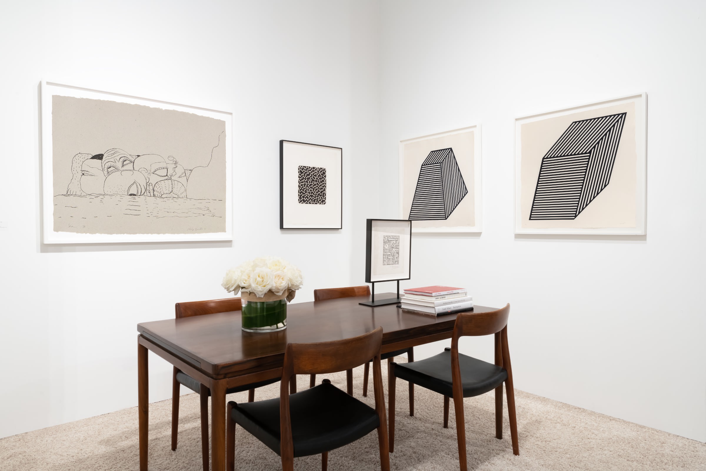 Closer installation view at Art Miami 2022 in Miami, with framed works arranged across two corner walls above a table with chairs, flowers, and books.