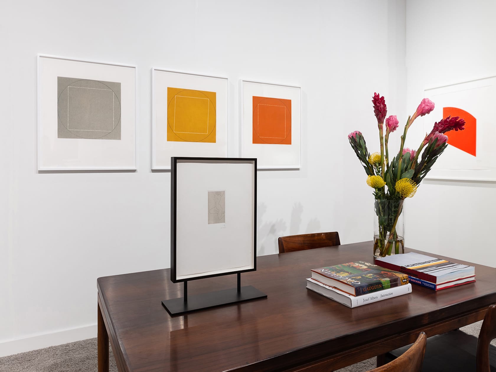 Closer installation view at Palm Beach Modern + Contemporary 2022 in West Palm Beach, with framed works above a table with chairs, books, and flowers.