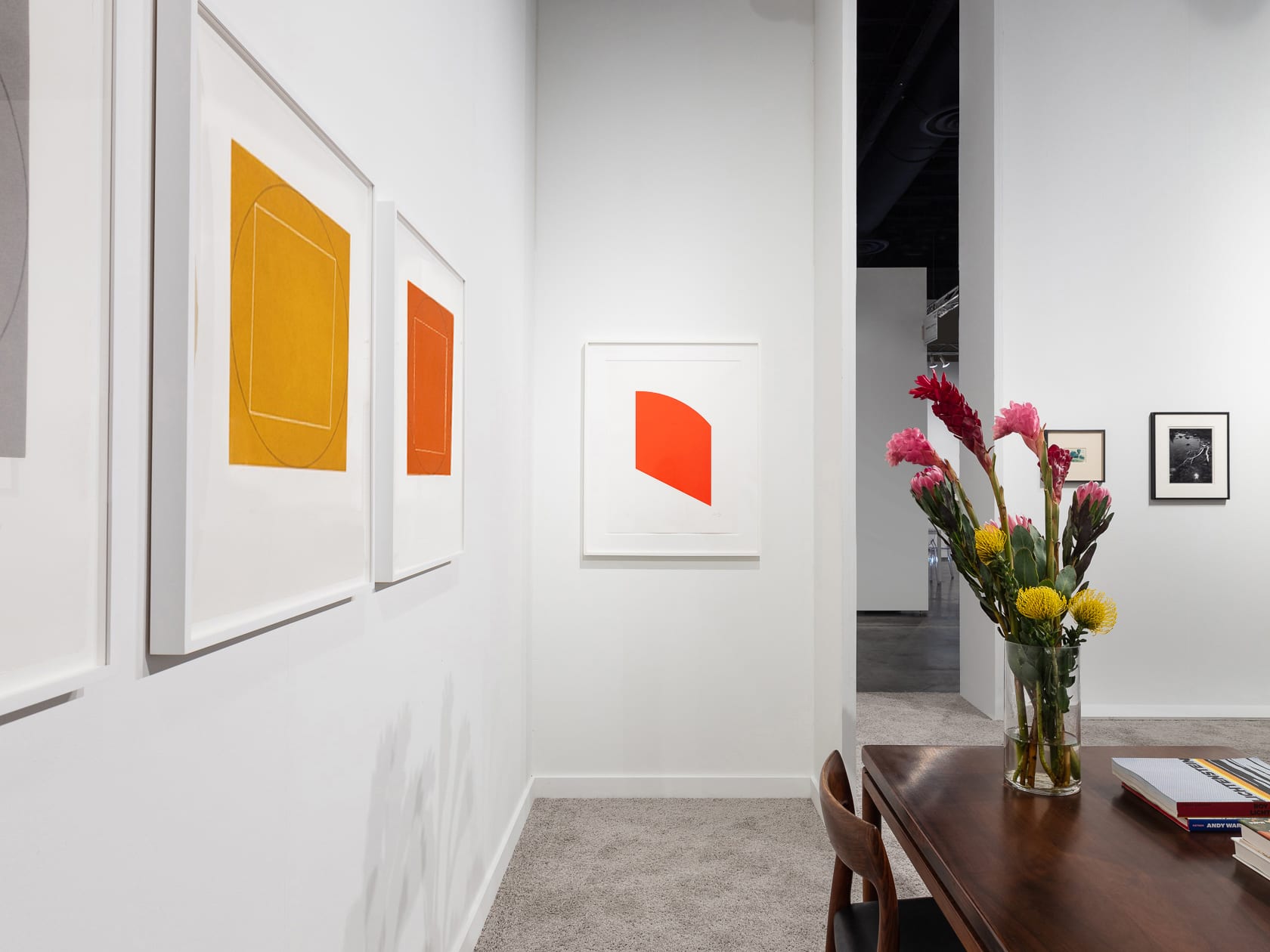 Installation view at Palm Beach Modern + Contemporary 2022 in West Palm Beach, with framed works above a table with flowers and a sightline through an opening into another booth section.