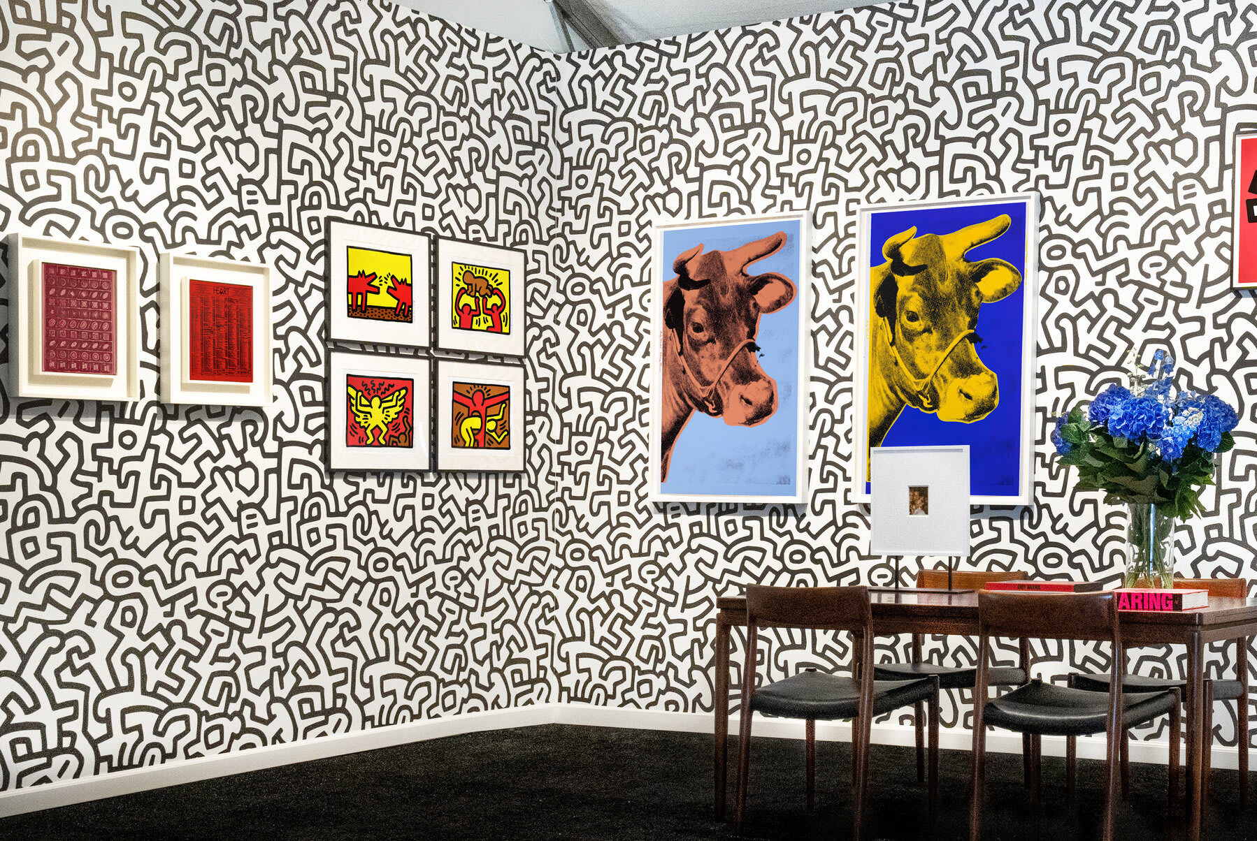 Installation view at Hamptons Fine Art Fair 2022 in Southampton, with framed works across two corner walls above a table with chairs and flowers.