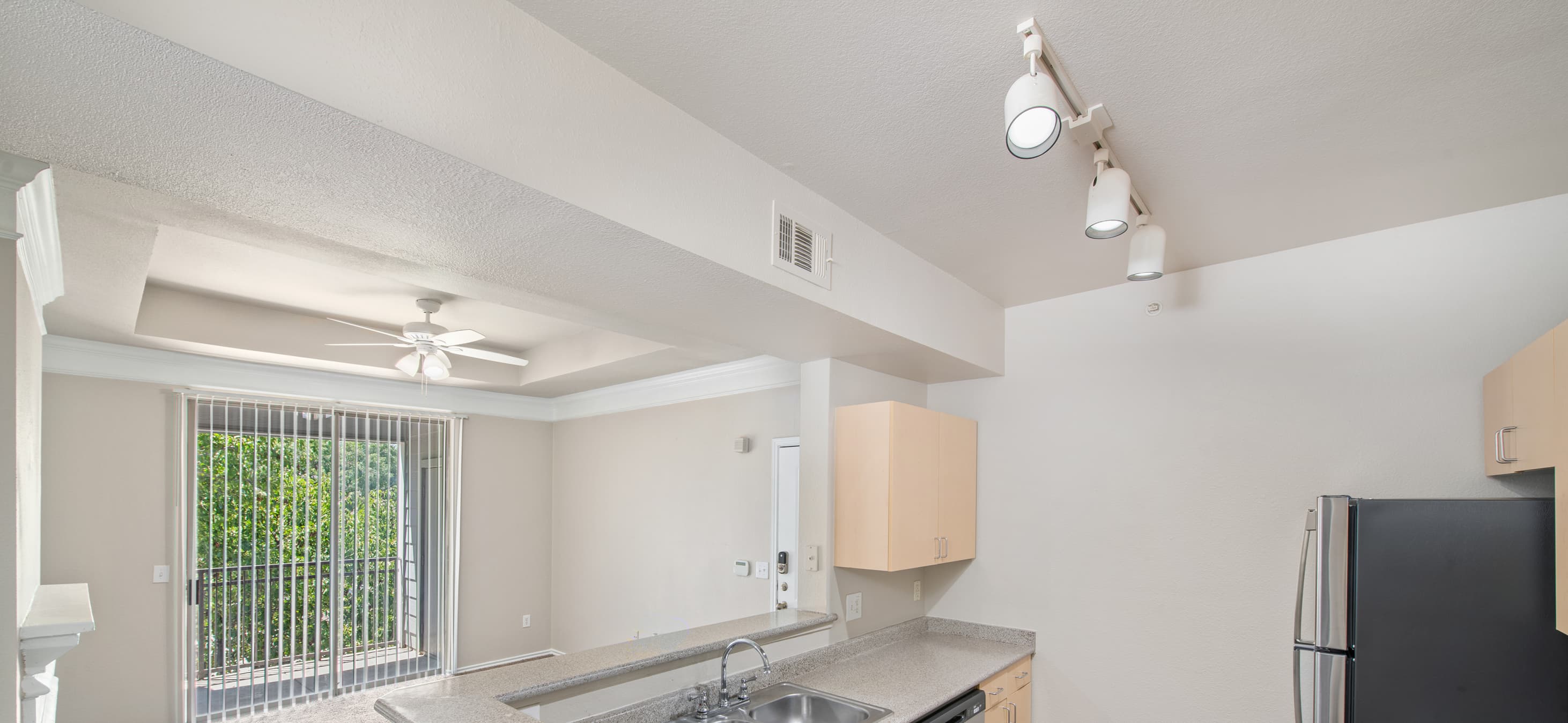 An outward view from kitchen features track ceiling lights over bar top counter and light tan wood cabinets positioned next to the living room with sliding glass doors at MAA Grand Courtyards luxury apartments in Grand Prairie, TX
