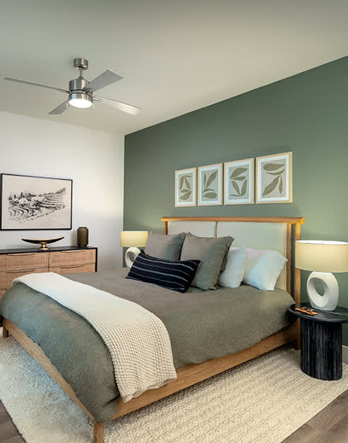 Bedroom with a ceiling fan, green accent wall with framed art above wooden bed frame with an area rug on wood floors at MAA Nixie luxury apartments in Durham, NC