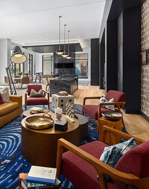 Modern clubhouse with multiple stylish lounge sitting areas, wall-mounted TV next to a communal kitchen bar with hanging light fixtures, and floor length picture windows at MAA Nixie luxury apartments in Durham, NC