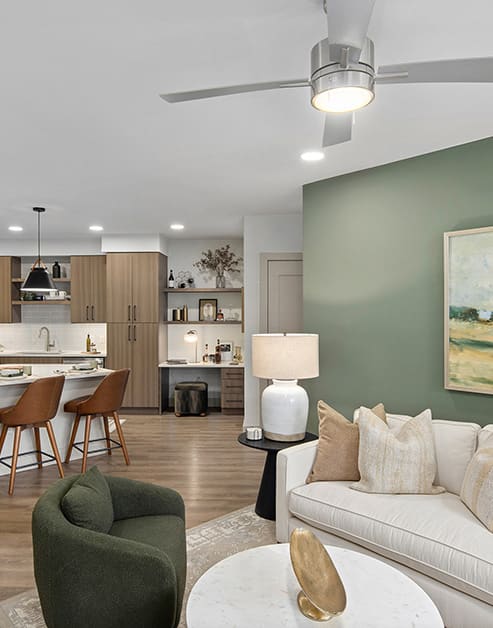 Model living room with a ceiling fan, a large wall painting on green accent wall with a white sofa and dark green armchairs next to a small white round coffee table on wood floors next to kitchen at MAA Nixie luxury apartments in Durham, NC