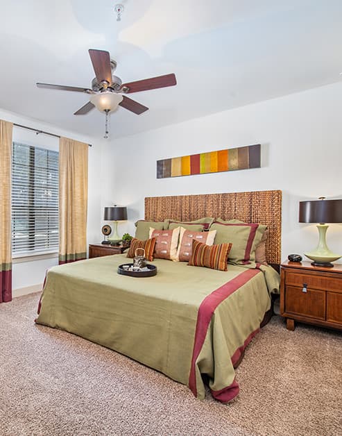 Carpeted bedroom with a ceiling fan, wall art above bed with accent pillows, night stands with lamps next to a large window with blinds and curtains at MAA Westover Hills luxury apartments in San Antonio, TX