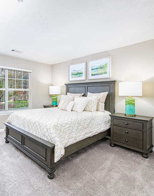 Model bedroom with wooden bed set with nightstands, next to a window with horizontal blinds at MAA Berkeley Lake luxury apartments in Duluth, GA