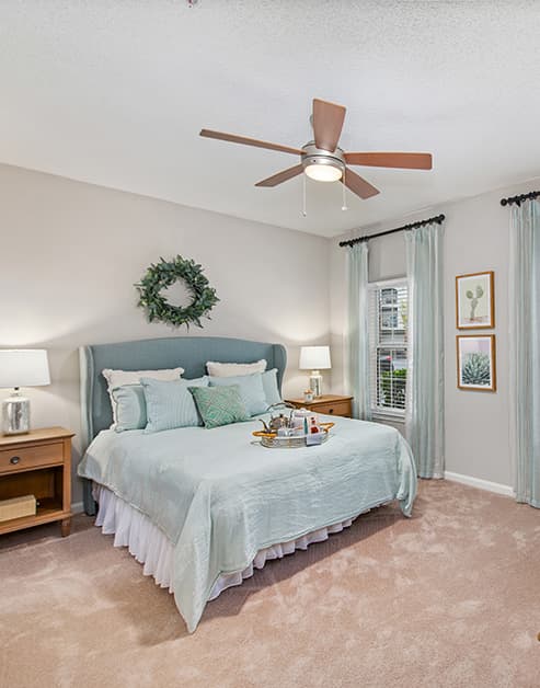 Model bedroom with ceiling fan and wall paintings, queen bed with wood night stands next to a window with blinds at MAA South Park luxury apartments in Charlotte, NC