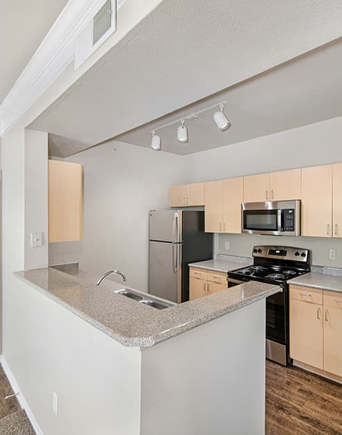 A modern kitchen bar top counter and track ceiling lights with light tan wood cabinets and stainless steel appliances at MAA Grand Courtyards luxury apartments in Grand Prairie, TX