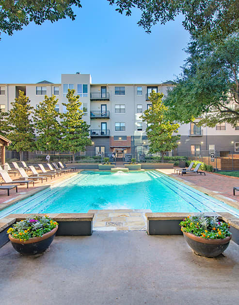 A rectangular swimming pool surrounded by lounge chairs, potted plants and tall trees at MAA Heights luxury apartments in Dallas, TX