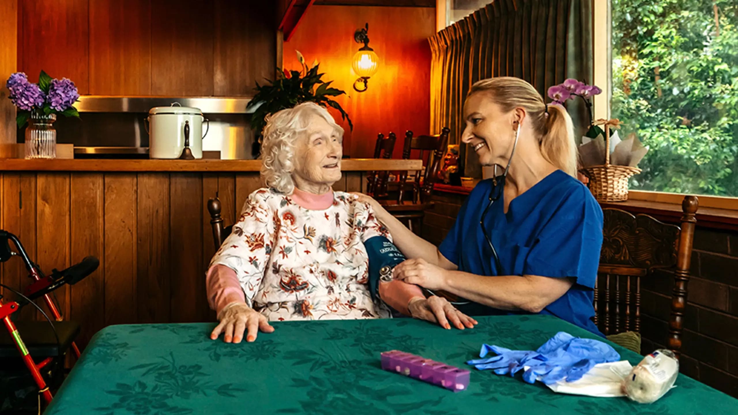 Registered Nurse takes a blood pressure reading for their older client at their dining table. 