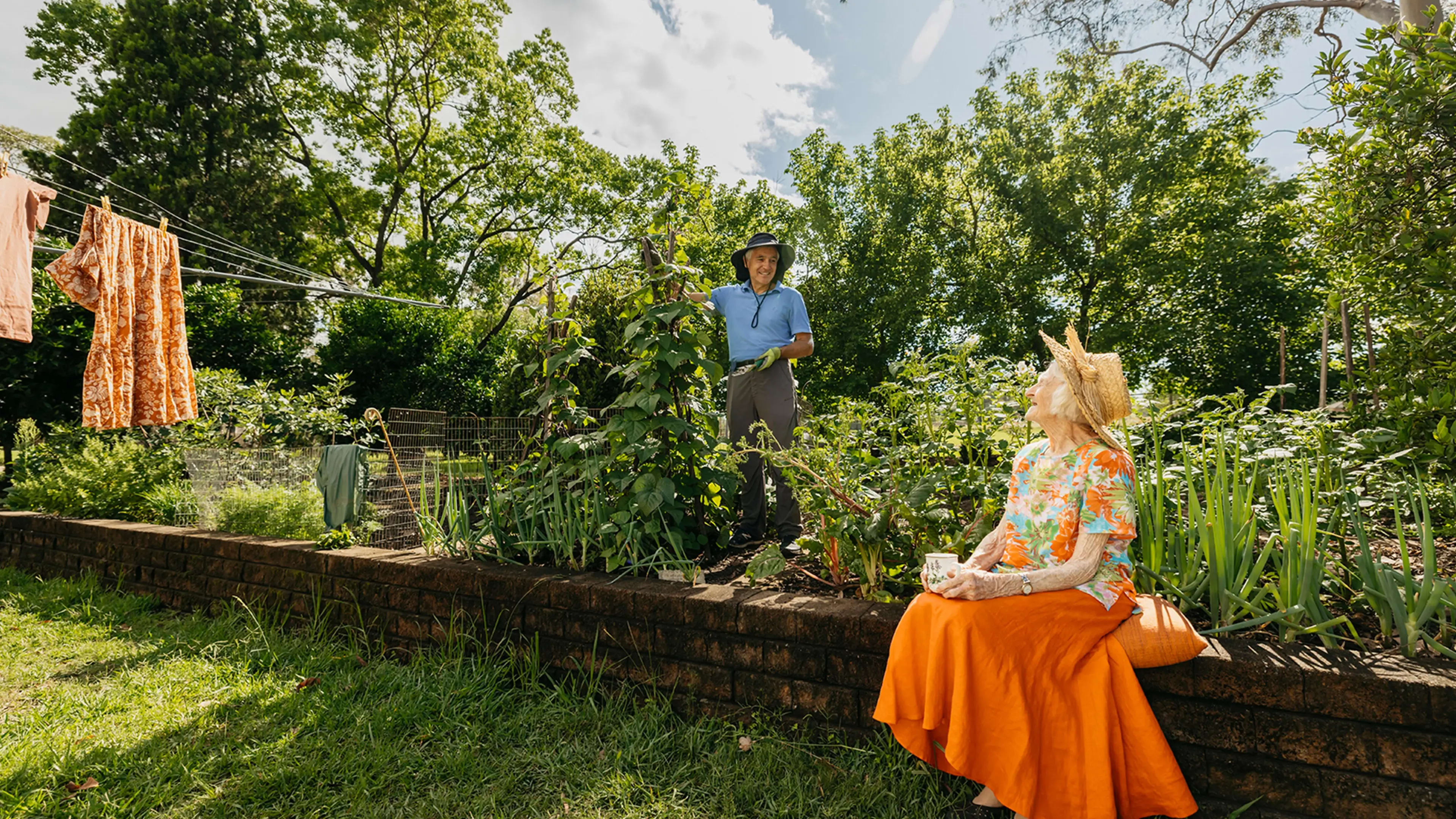 Support worker prunes plant in garden while chatting to their older client sitting on garden wall.