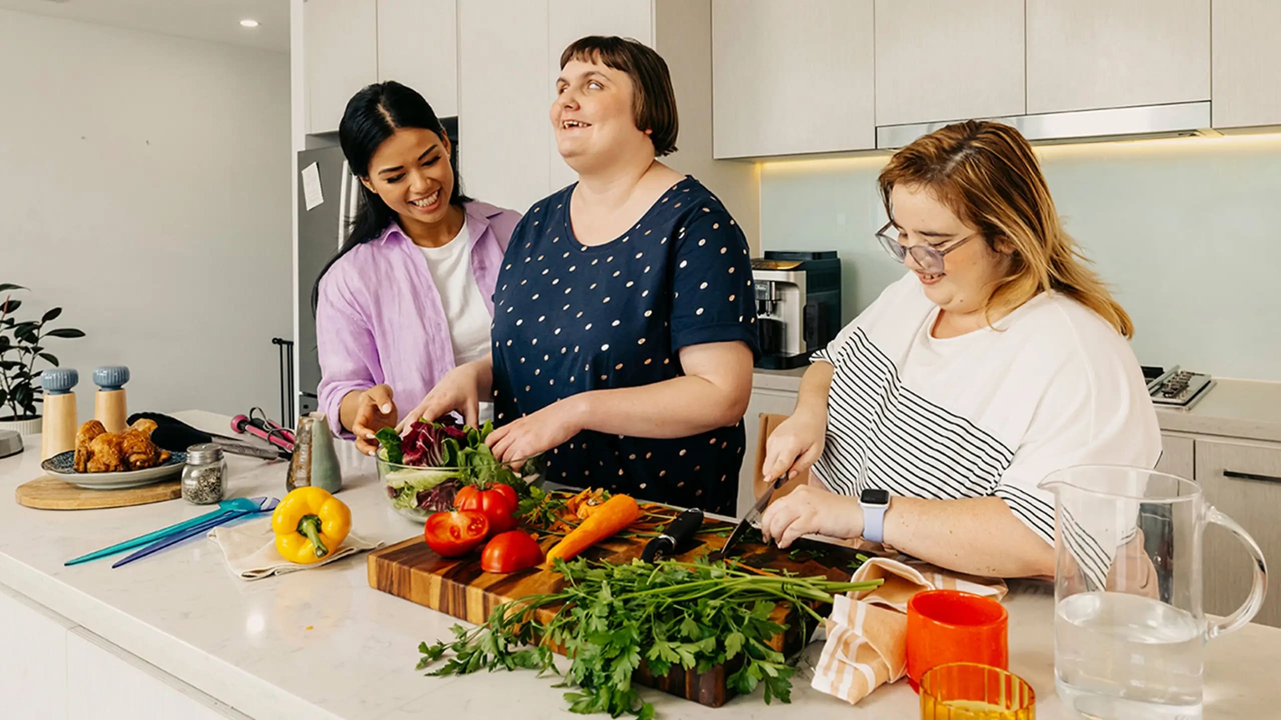 Support worker helps two people with disabilities prepare vegetables at the kitchen bench.