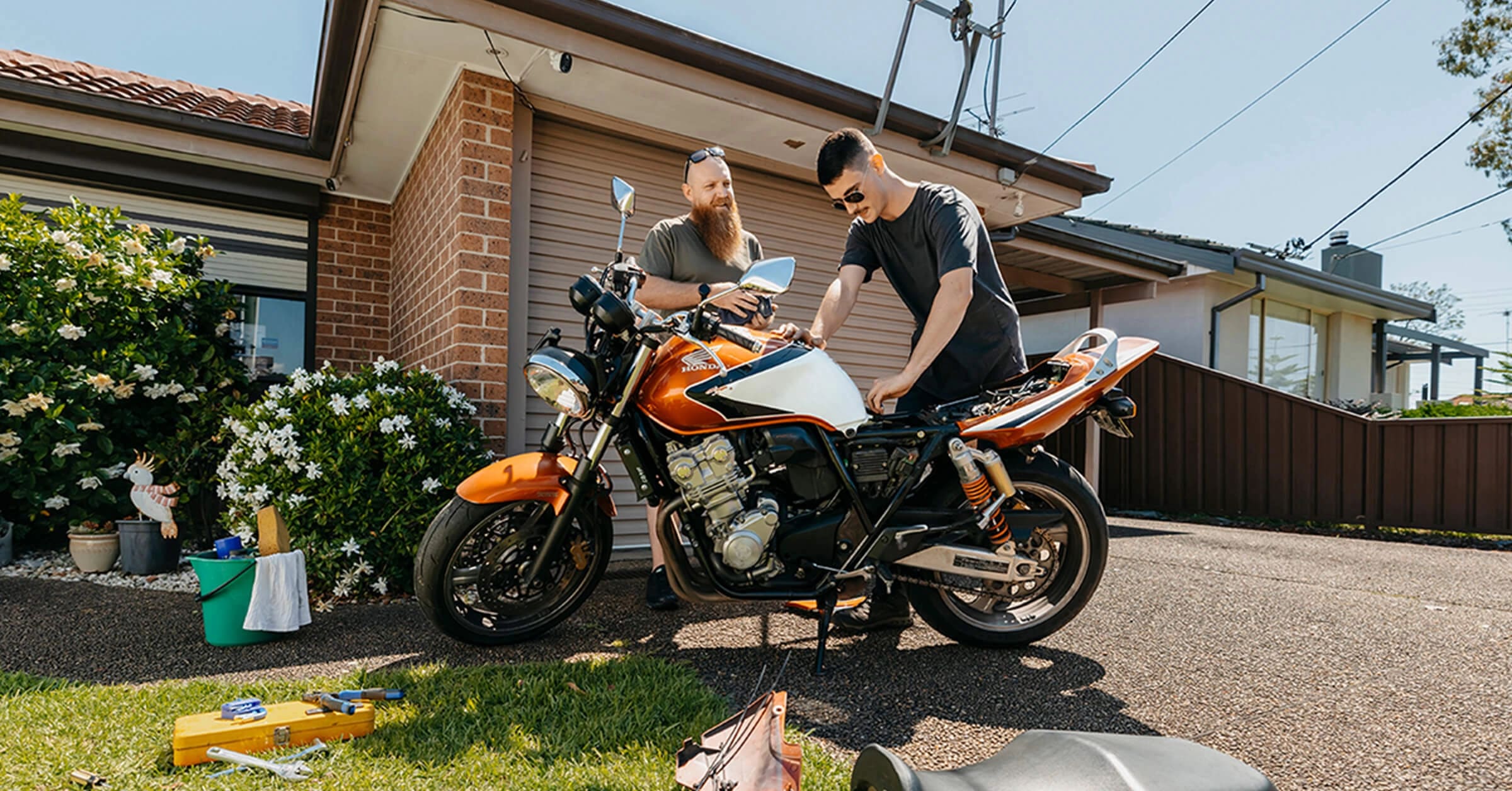 Support worker and young adult with autism and Tourette syndrome work on a motorbike in sunny driveway.